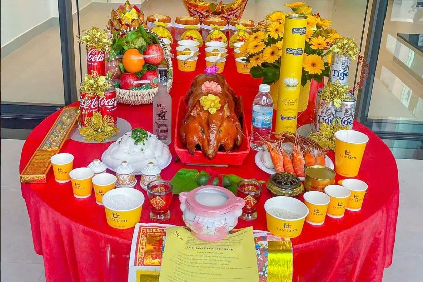 Mâm cúng gồm heo quay, tôm, giỏ trái cây, hoa đồng tiền và nhang đèn trên bàn tròn phủ khăn đỏ