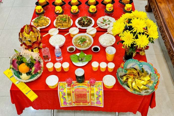 Mâm cơm cúng với cơm trắng, canh, các món chay, xôi, trái cây và hoa cúc vàng bày trên bàn phủ khăn đỏ