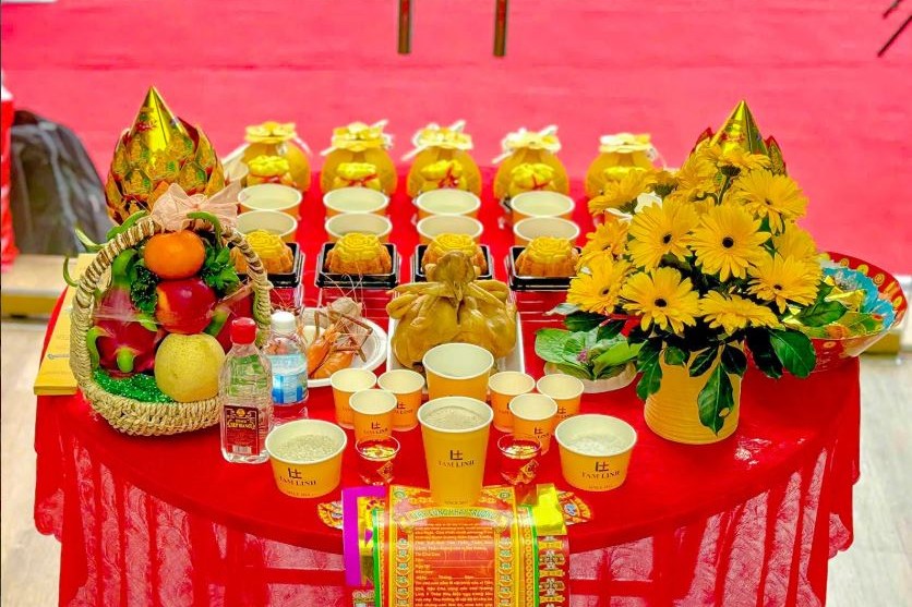 Mâm cúng khai trương với gà luộc, bánh bao túi tiền, xôi chè, giỏ trái cây và hoa đồng tiền vàng đặt trước cửa hàng