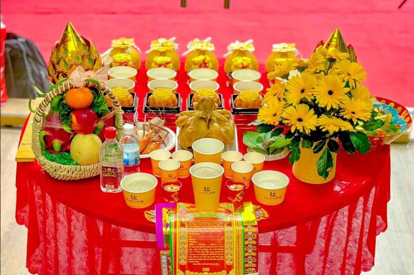 Mâm cúng với gà luộc, xôi, chè, bánh bao túi tiền, giỏ trái cây và hoa đồng tiền vàng bày trước cửa hàng