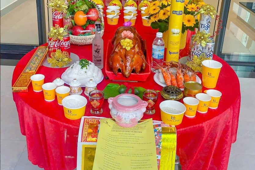 Mâm cúng với heo quay, tôm, trái cây, hoa đồng tiền và nhang đèn bày trên bàn tròn phủ khăn đỏ trong ngôi nhà mới xây