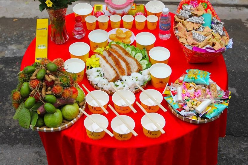 Mâm cúng cô hồn ngoài trời với heo quay, bánh hỏi, cháo trắng, trái cây, bánh kẹo, hoa cúc và giấy tiền trên bàn tròn phủ khăn đỏ